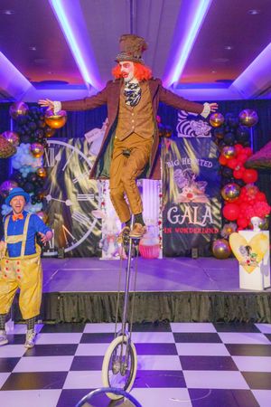 A performer in a brown costume balances on a unicycle on a checkered stage with purple lighting and festive decorations.