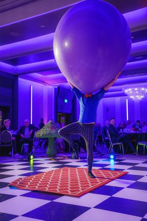 A large blue balloon on a stand illuminated by purple lighting in a ballroom with a checkered floor and red carpet.