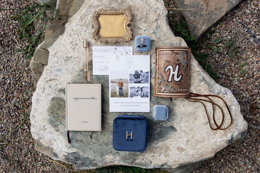 Wedding stationery and accessories arranged on a grey stone surface including invitations and a monogrammed leather item.