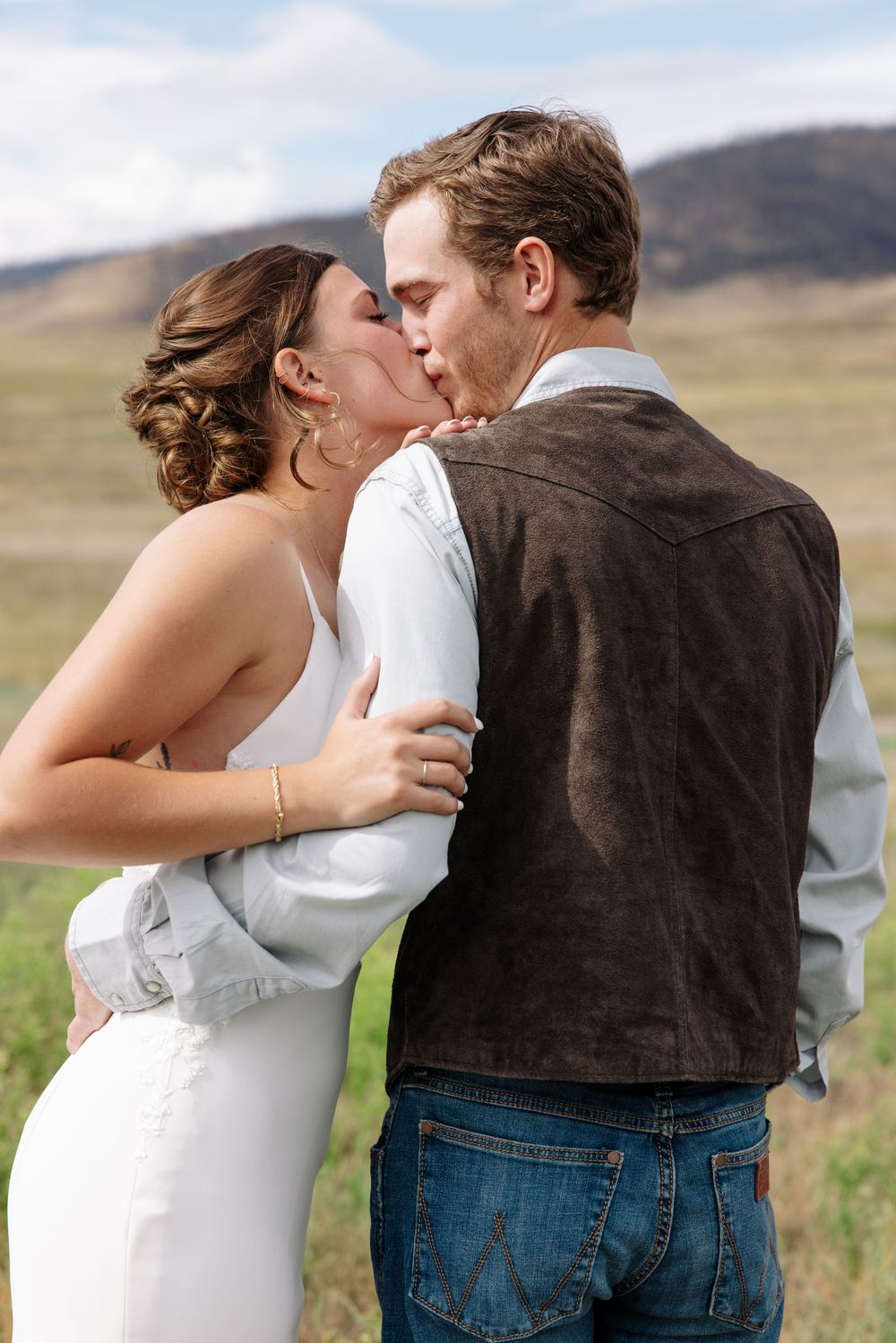 A couple shares a romantic kiss in a field with rolling hills in the background on their wedding day.