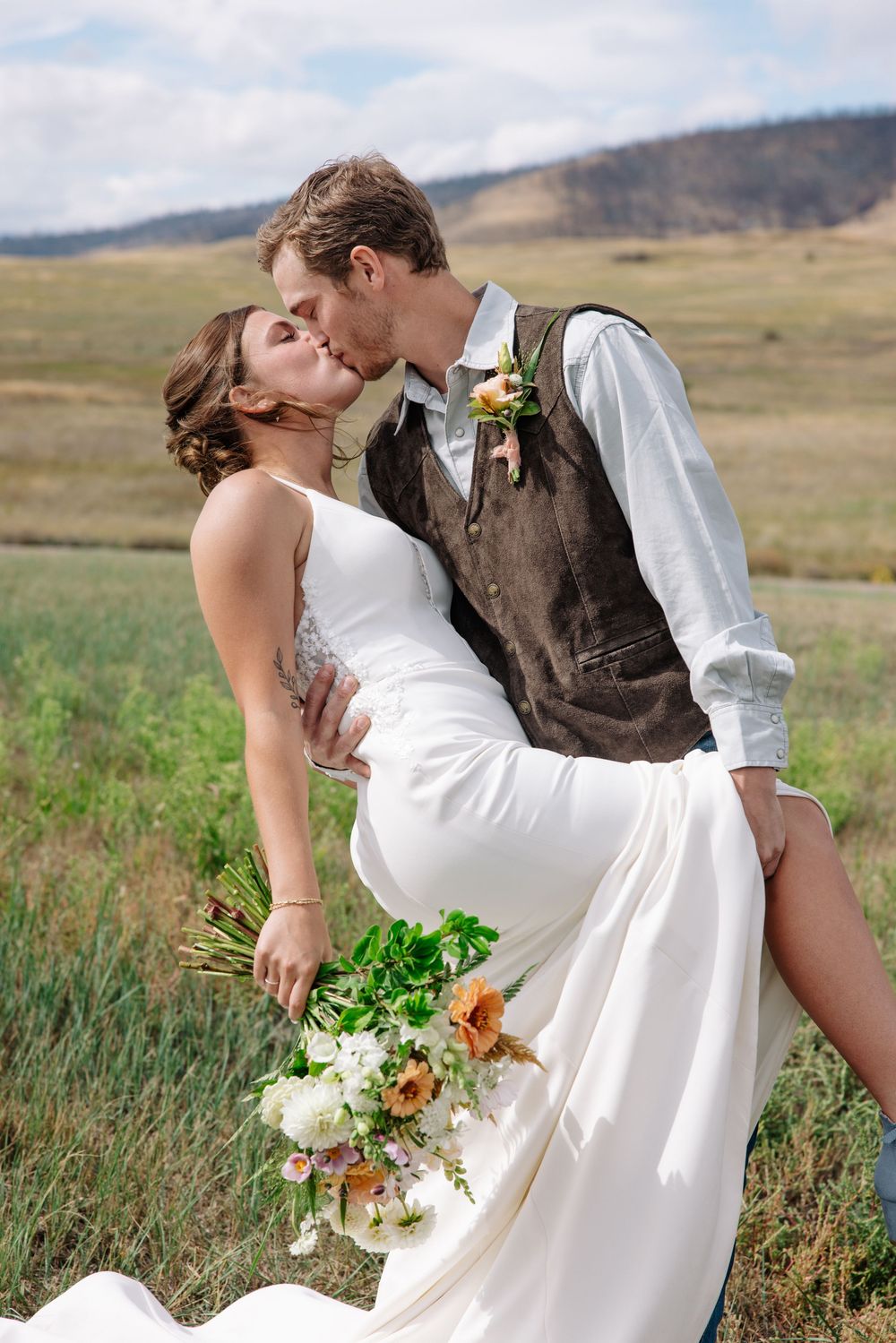 A romantic wedding moment with a dip kiss pose in a scenic mountain meadow with brown hills in the background.