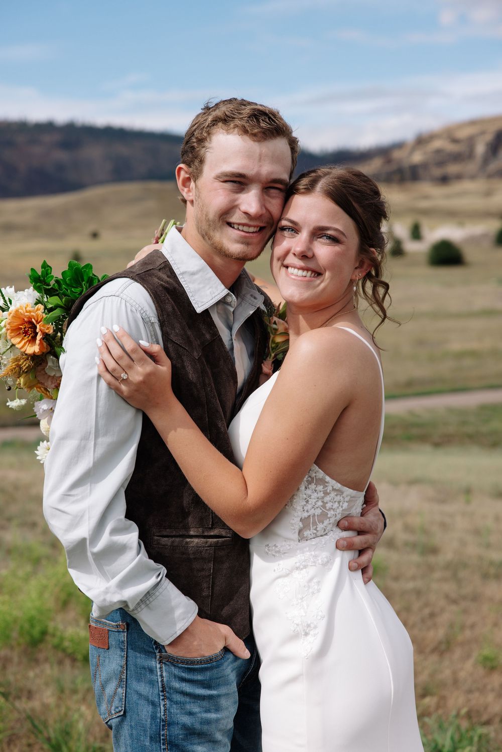 A happy newlywed couple embraces outdoors with mountains and fields in the background.