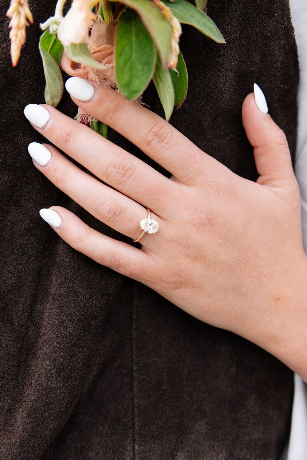 Close-up of an elegant engagement ring with white polished nails against dark fabric and delicate floral details.