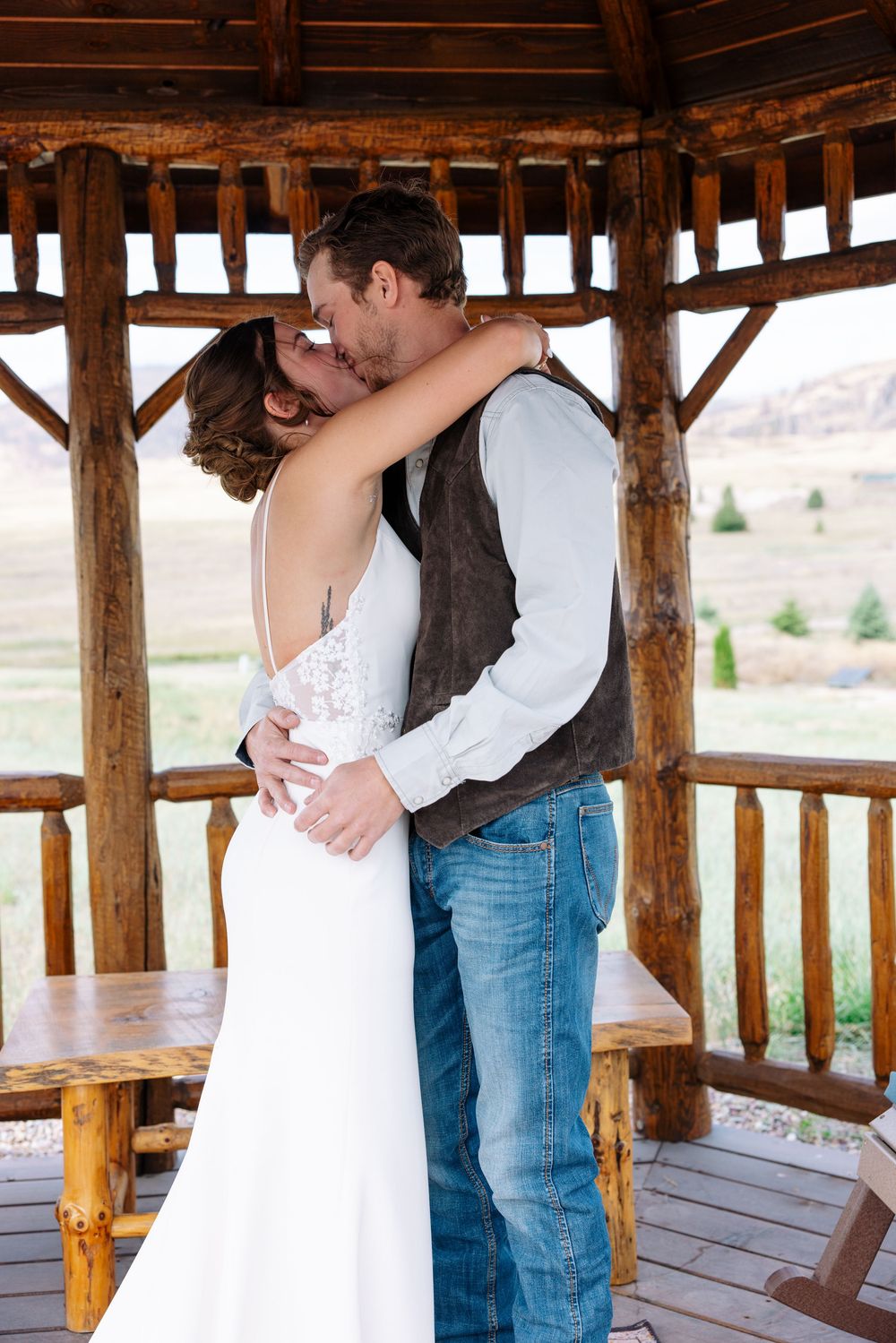 Romantic couple embrace in rustic wooden gazebo with mountain landscape behind, bride in white dress and groom in vest and jeans.