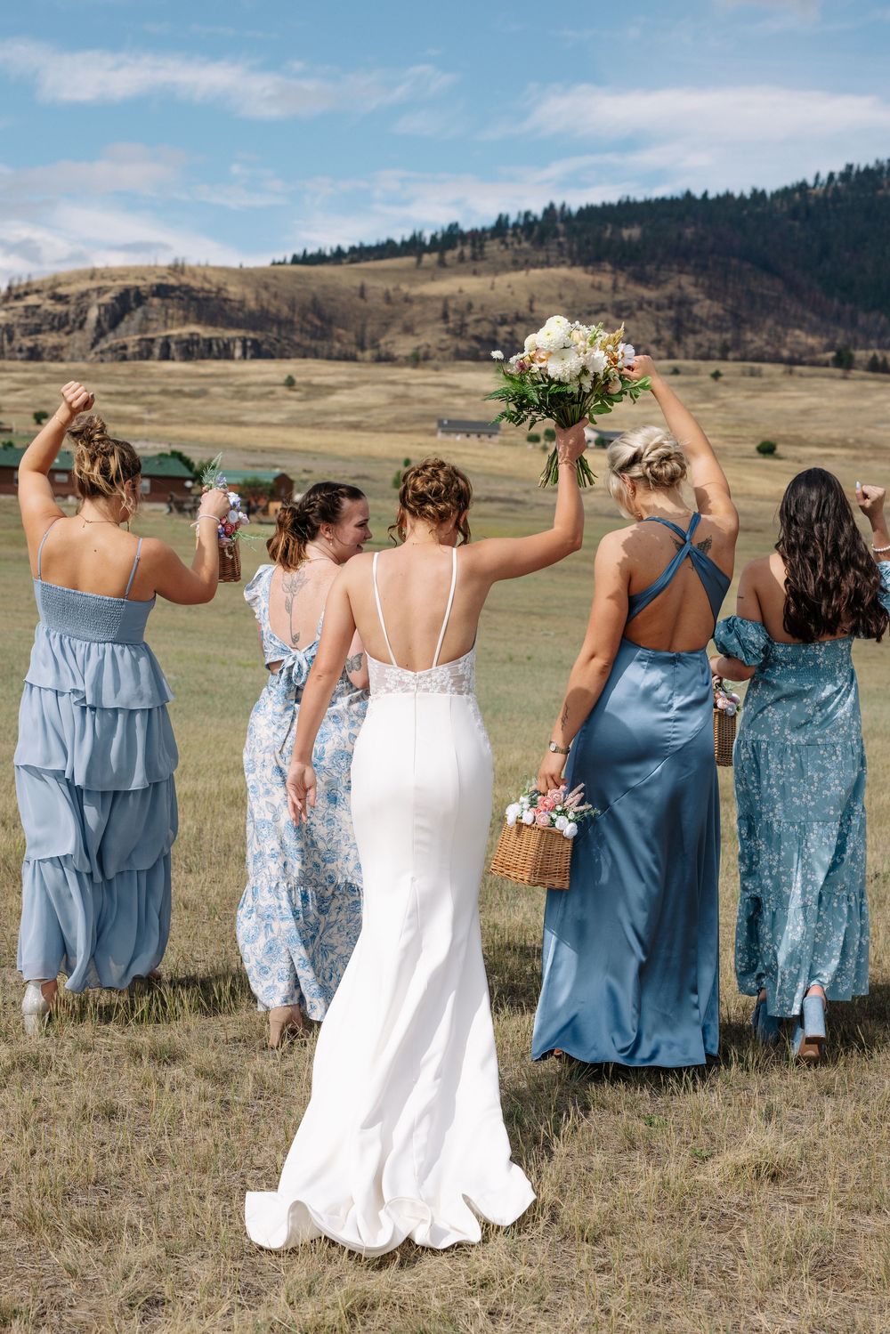 A bridal party in blue and floral dresses celebrating on a hillside with mountain views in the background.
