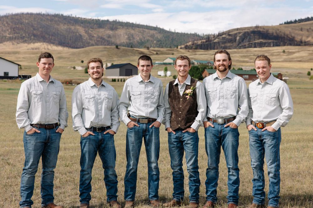 A group of six men stand together in a row outdoors wearing light colored shirts, jeans and boots against a rural hillside.