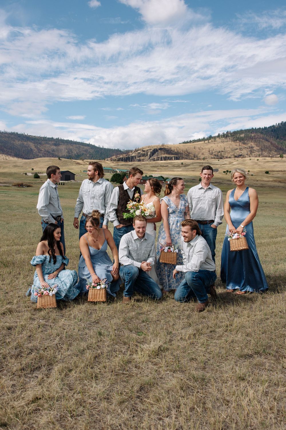 A wedding party poses together in a scenic mountain valley field with picnic baskets on a sunny day.