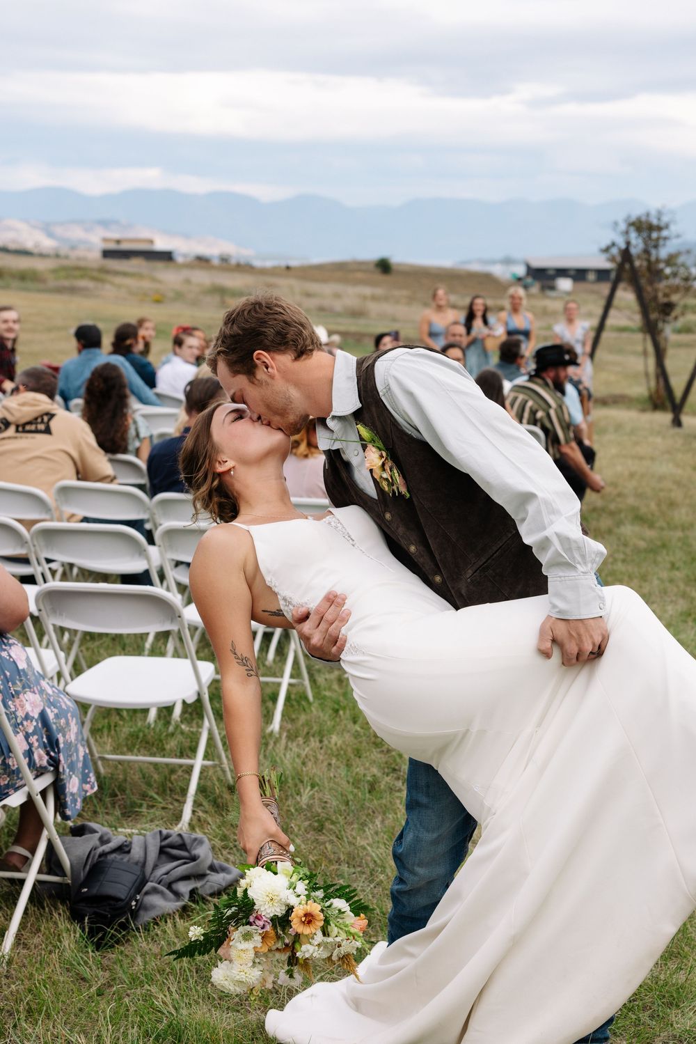 A romantic wedding kiss at an outdoor ceremony with mountain views, folding chairs, and a bouquet of flowers.
