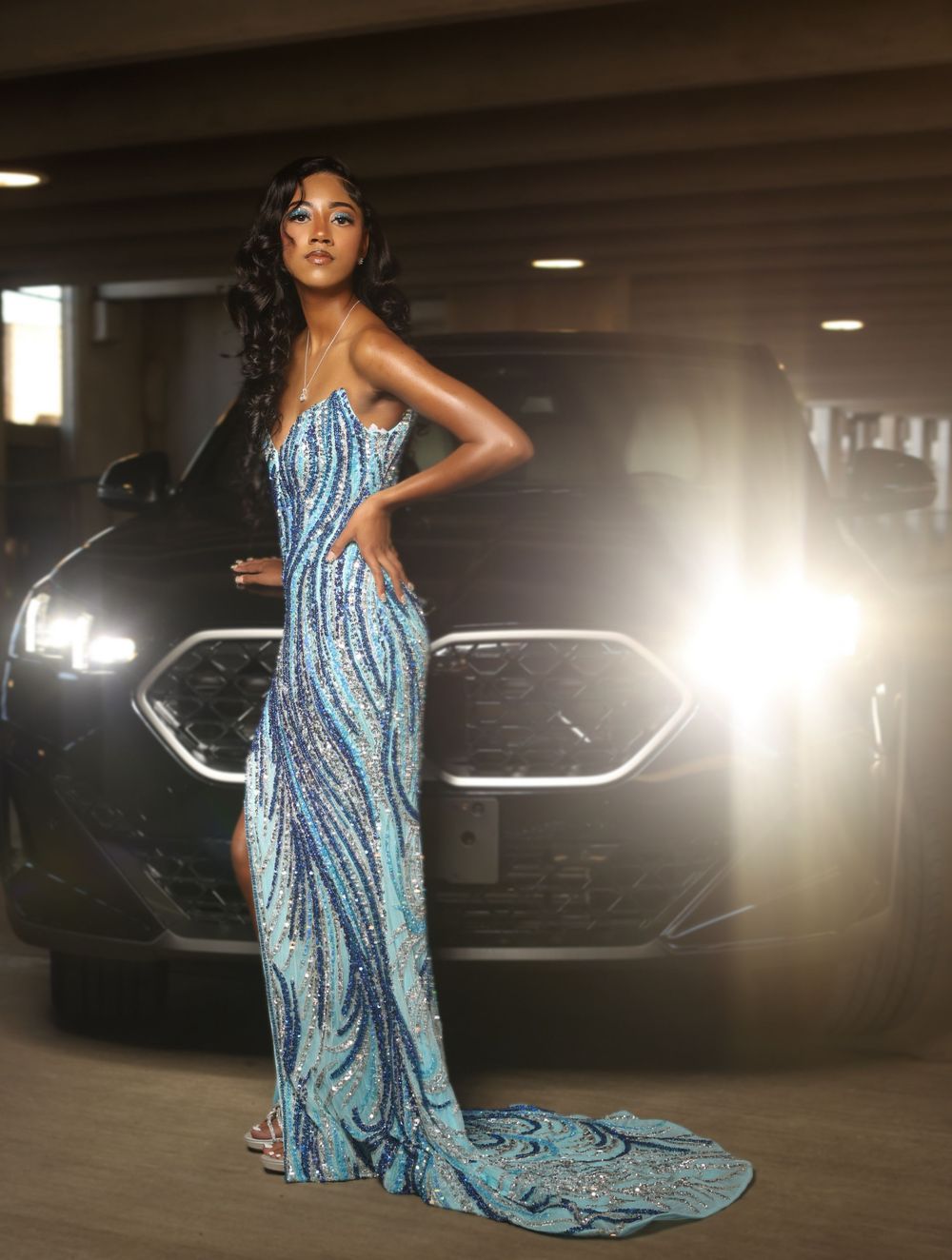 Elegant woman in stunning blue sequined gown poses dramatically in front of luxury car at night.