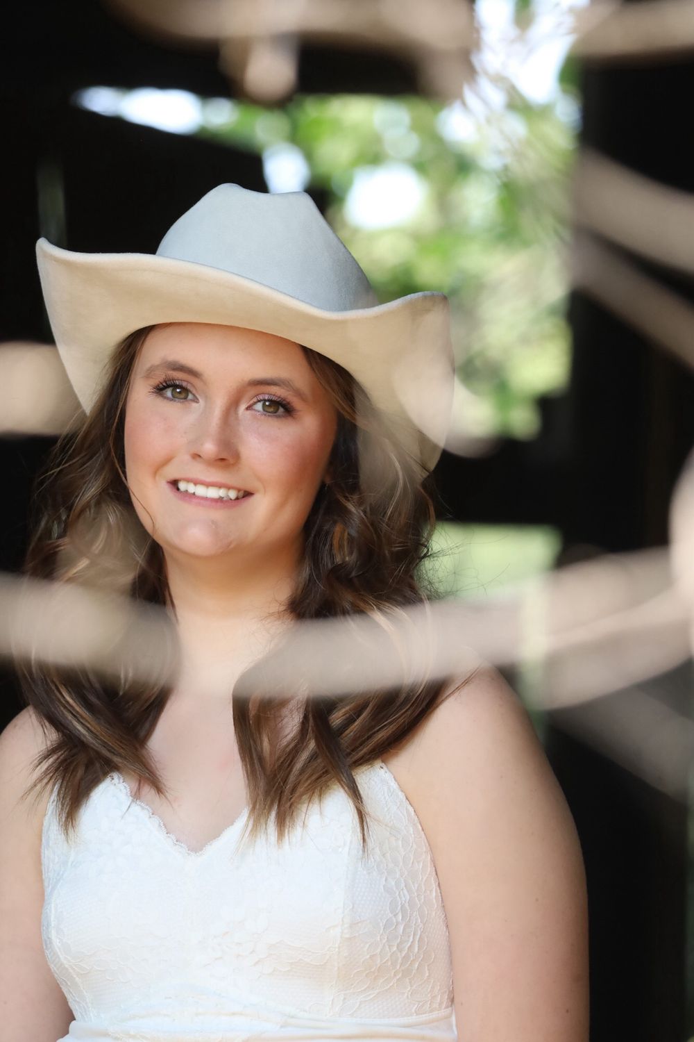 Smiling young woman in white dress and cowboy hat poses in rustic barn setting outdoors.