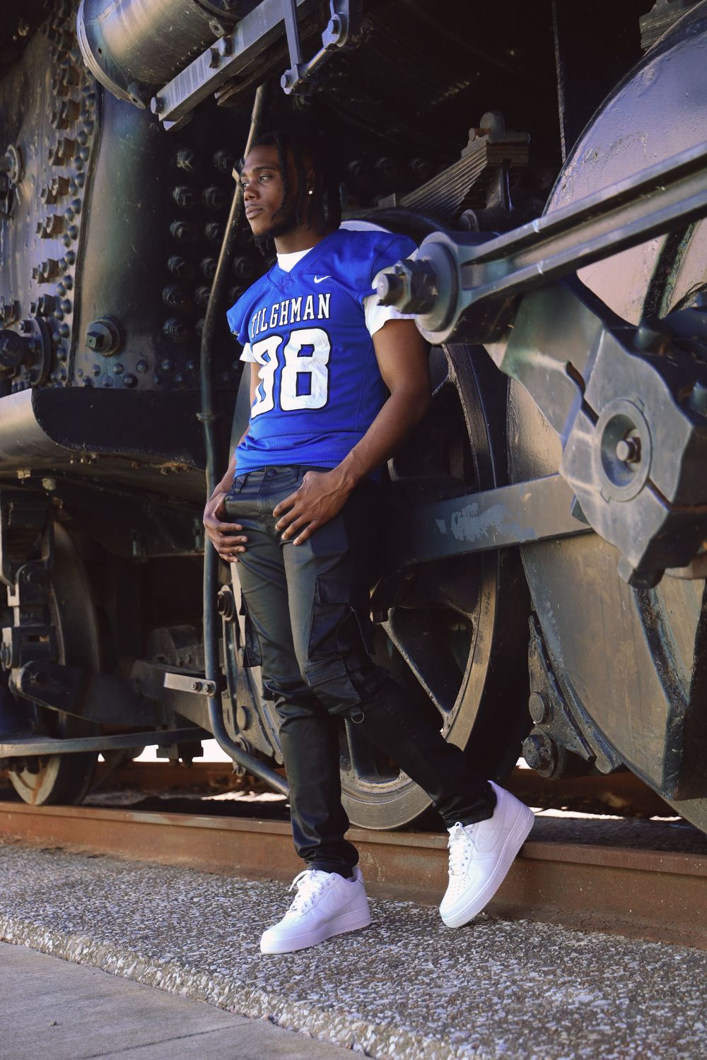 Young athlete in blue football jersey number 38 poses beside vintage locomotive on railroad tracks.