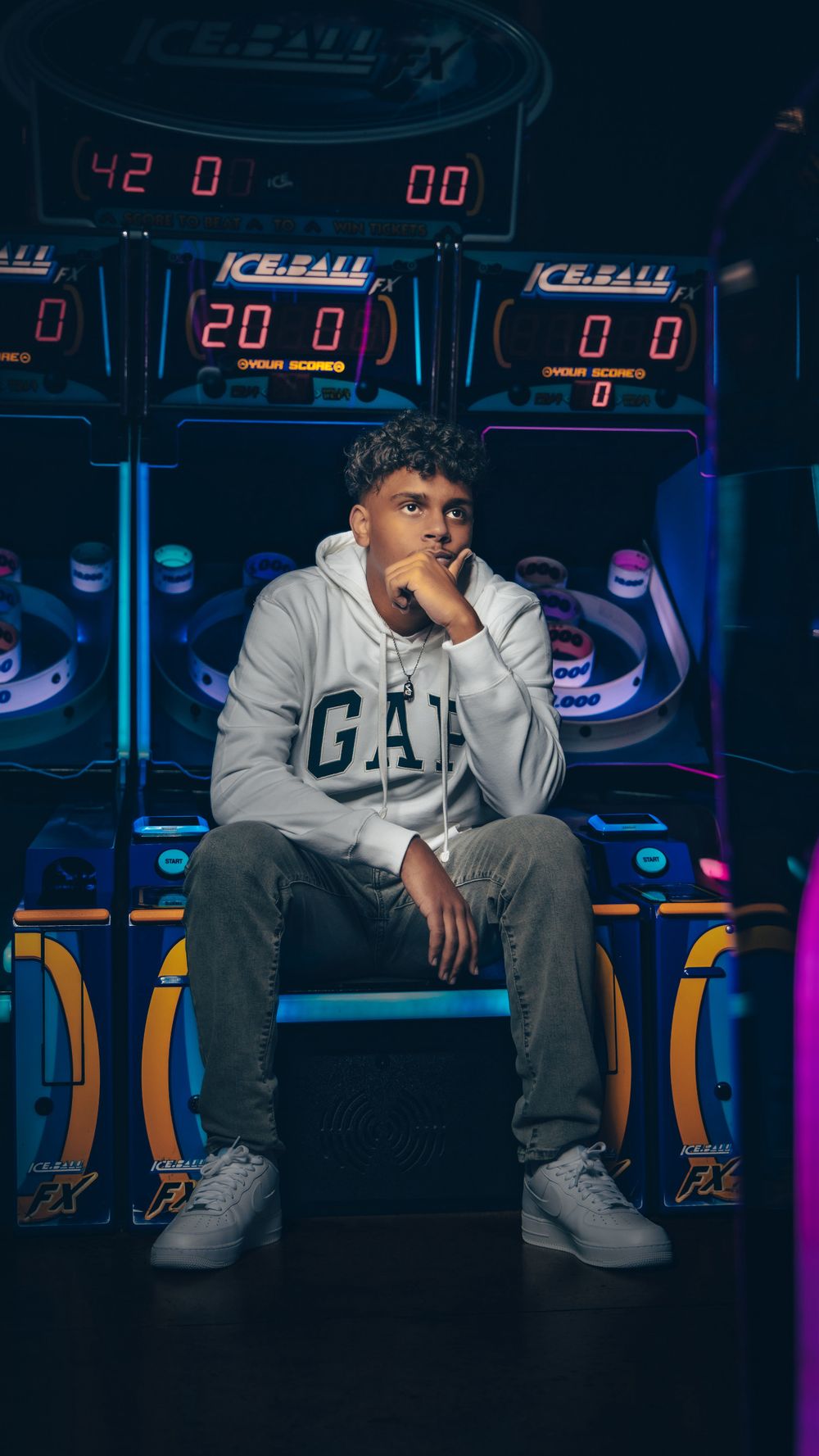 Young man in GAP hoodie sits pensively in front of colorful arcade Ice Ball machines.