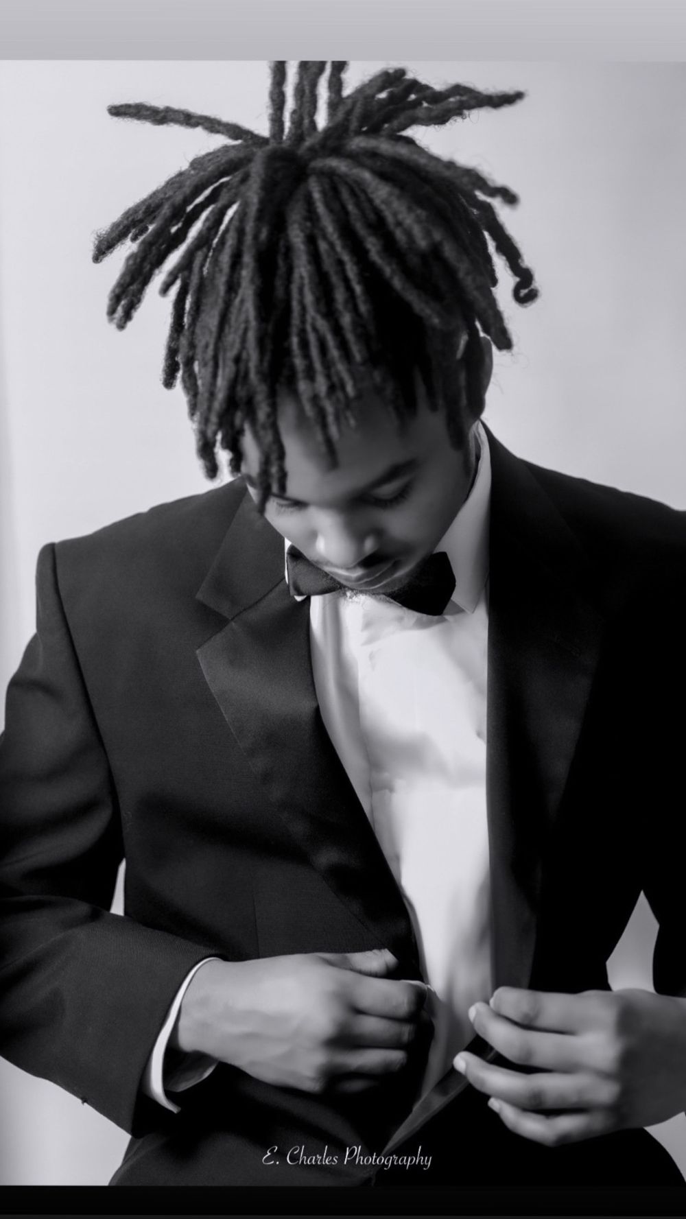 Elegant black and white portrait of young man with dreadlocks adjusting formal tuxedo and bow tie.