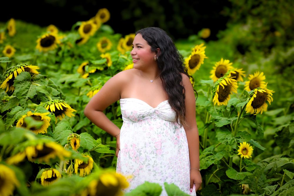 Young woman in white floral dress stands gracefully among tall sunflowers in a sunny field.