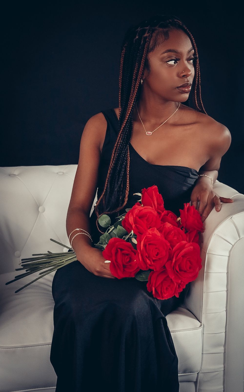 Young Black woman with long box braids holds red roses in a black strapless gown on a white sofa.