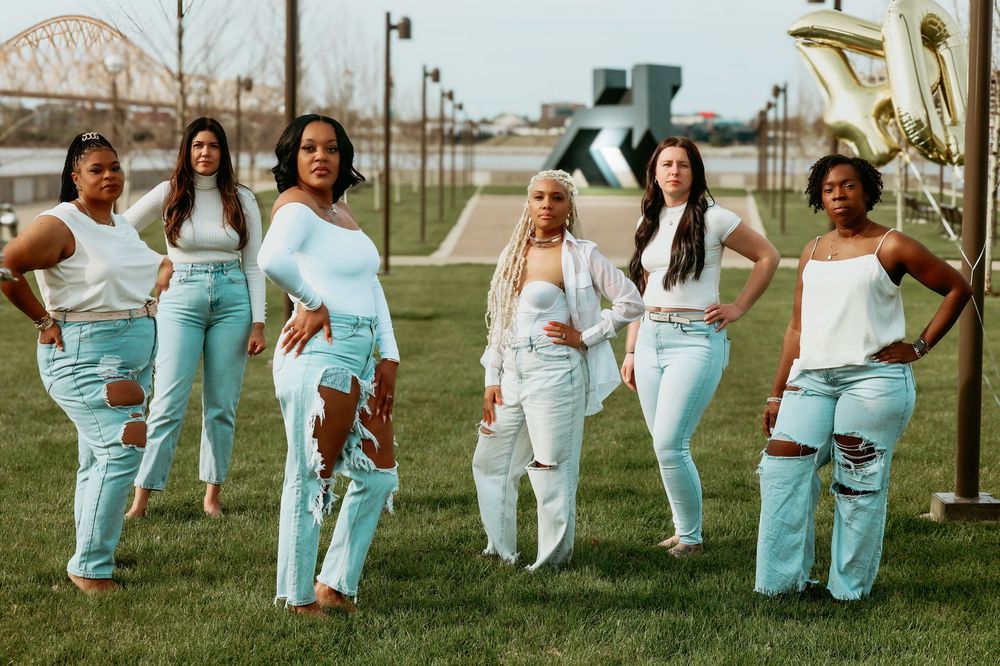 Six diverse women pose confidently in matching white tops and light blue ripped jeans outdoors by waterfront.
