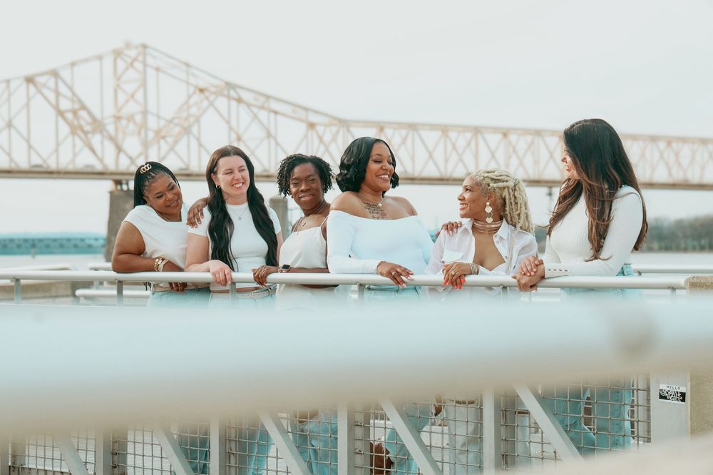 Six diverse women in white outfits smile together leaning on railing with bridge in background.