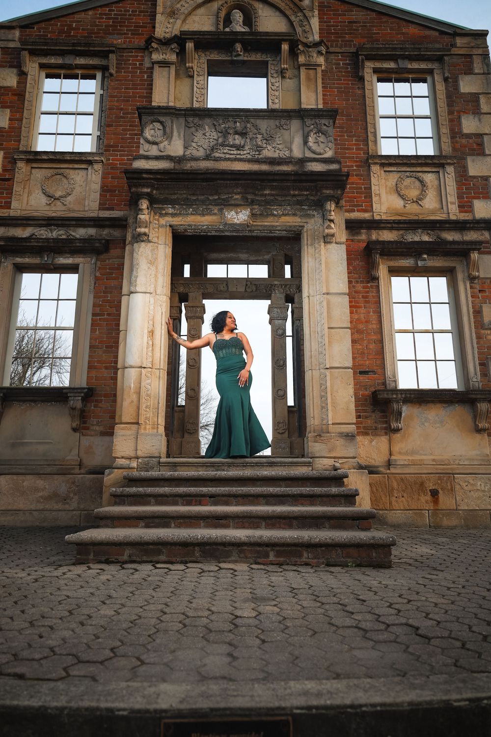 Woman in elegant emerald green gown poses in doorway of historic brick building with ornate architecture.