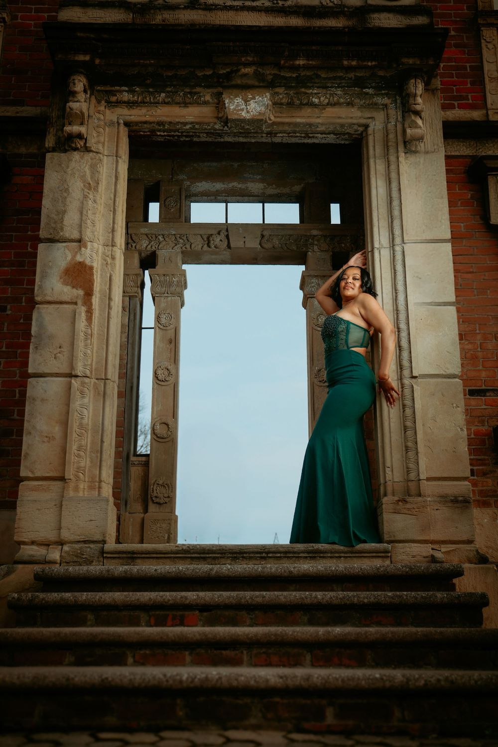 Woman in teal mermaid gown stands confidently in ornate stone archway against blue sky backdrop.