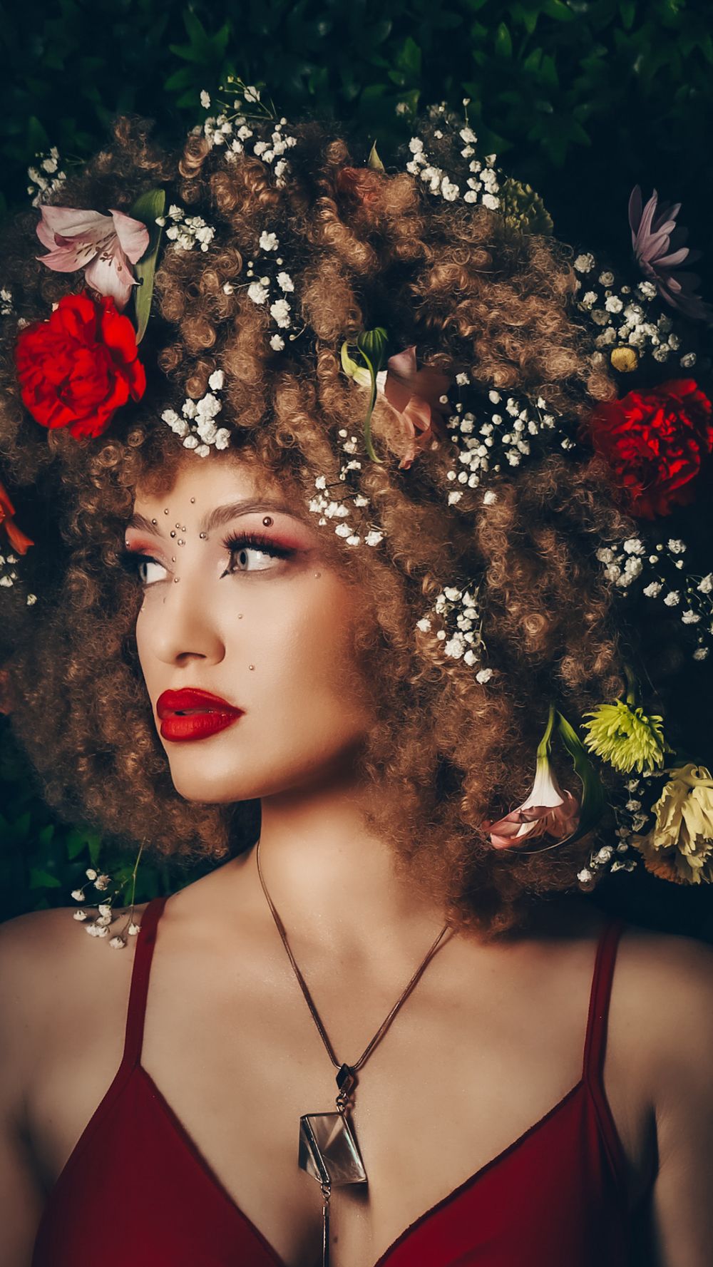 Portrait of woman with curly hair adorned with colorful flowers wearing red dress and crystal pendant.