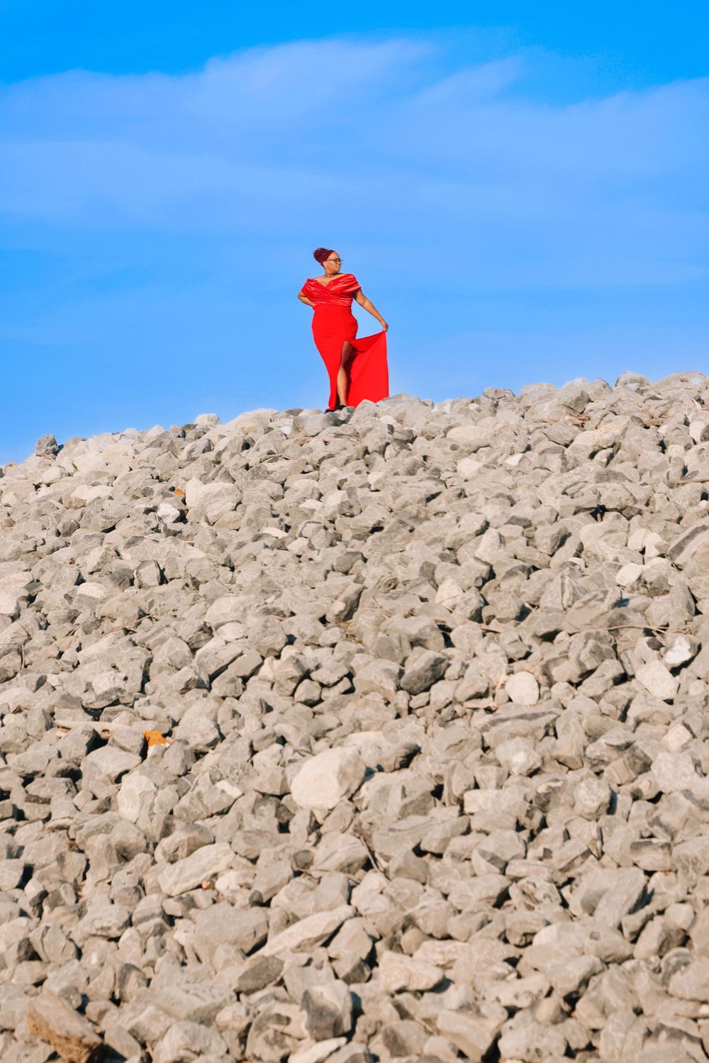 Woman in flowing red dress stands atop large pile of white rocks against bright blue sky.