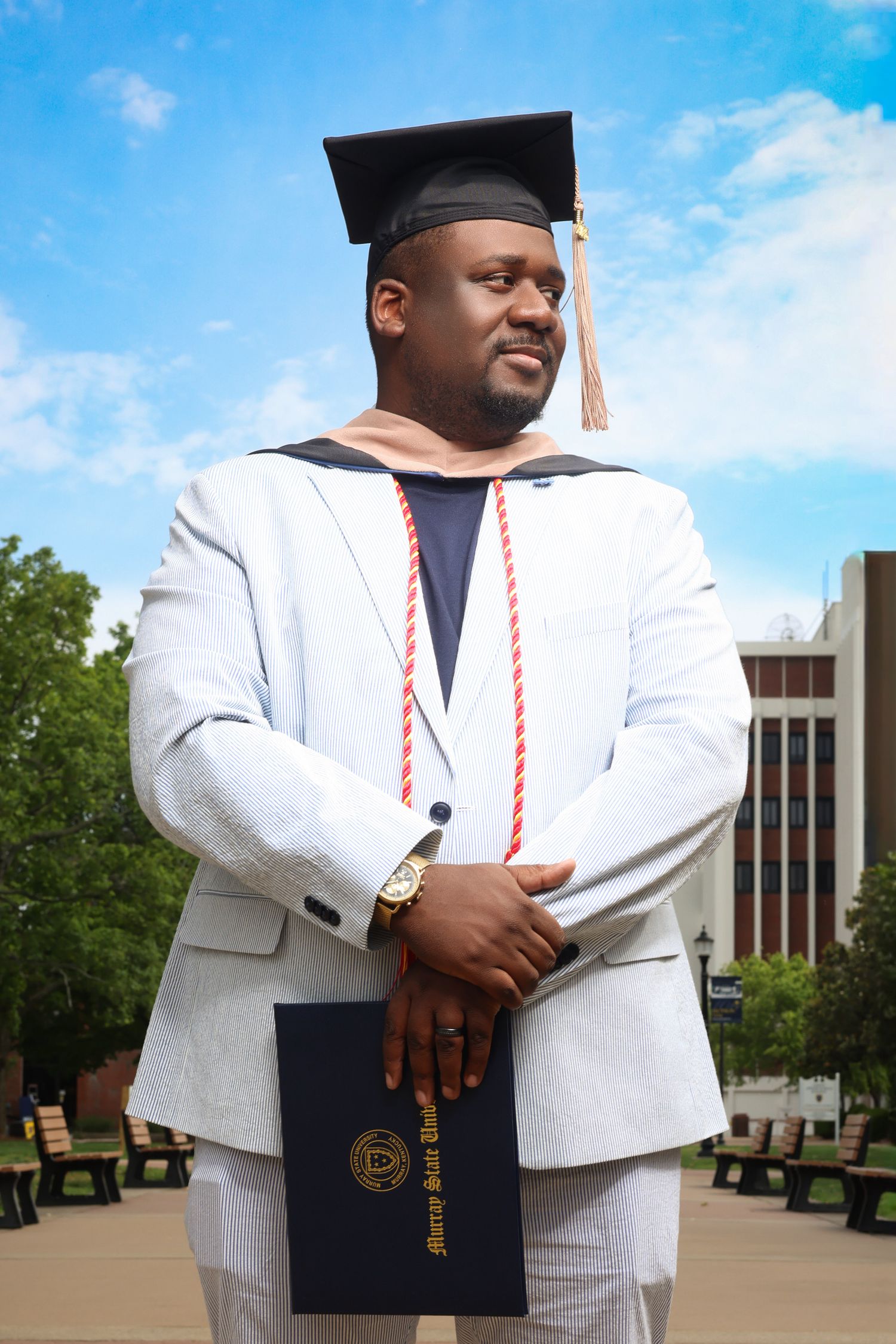 Graduate man in white suit and cap proudly holding diploma outdoors on a sunny university campus.