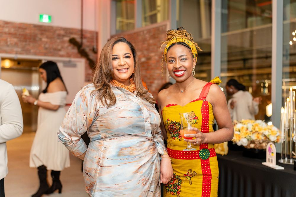 Two friends smiling at a social event, one wearing a metallic dress and one in vibrant yellow patterned attire.