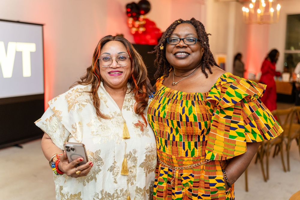 Two friends pose together at an event, one wearing a white dress and one in vibrant yellow African print attire.