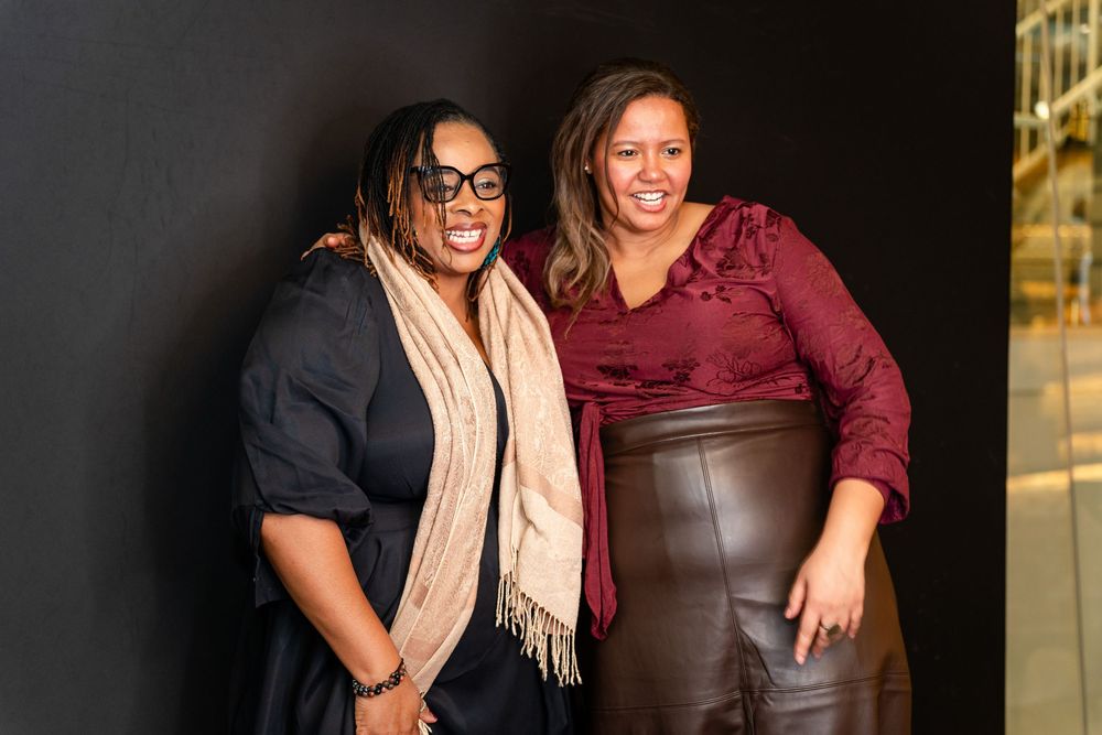 Two smiling women pose together against a dark background, one wearing glasses and a scarf, the other in maroon.