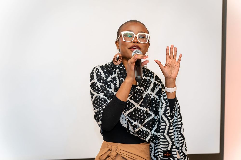 Speaker wearing a black and white geometric patterned sweater and glasses gestures while speaking into microphone during presentation.
