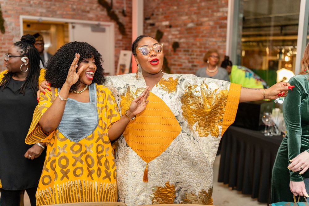 Two people in vibrant yellow and white African print outfits dance joyfully at an indoor celebration.