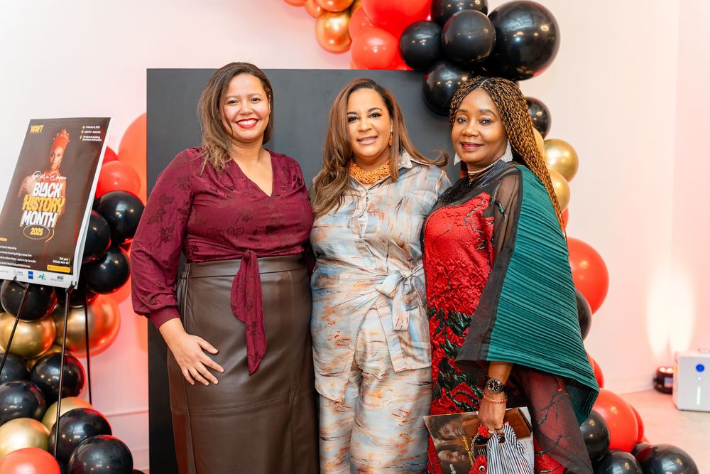 Three elegantly dressed women pose at a party with red and black balloon decorations in the background.