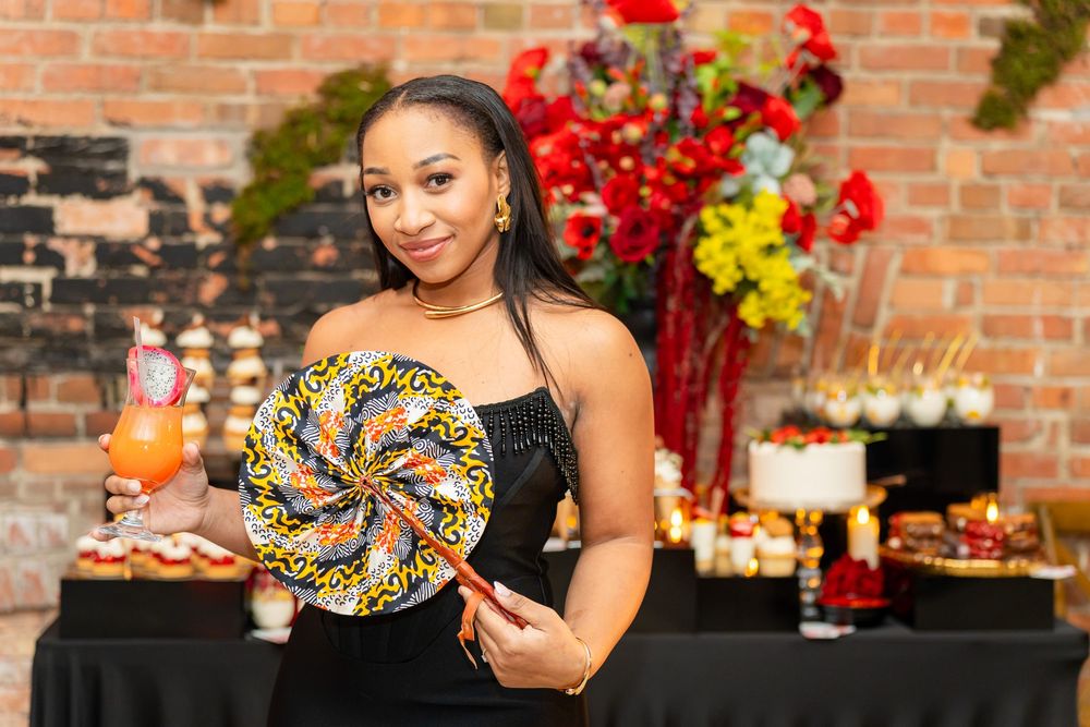 Someone holds a colorful lollipop decoration at a party with a festive dessert table and floral arrangements in the background.