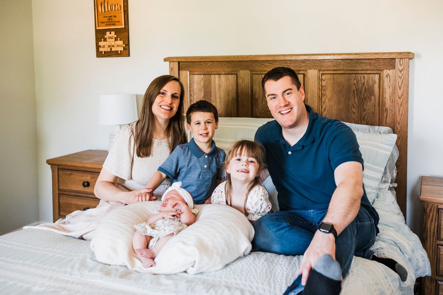 Family sits together on a bed with wooden headboard in a bright, airy bedroom interior.