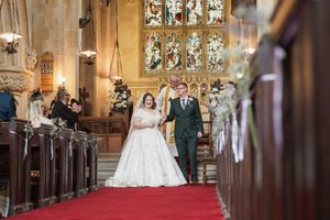 Wedding couple walks down red-carpeted church aisle with stained glass windows in background.