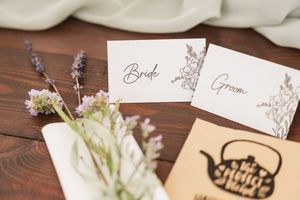 Wedding place cards with bride and groom text sit beside delicate lavender flowers on a wooden surface.