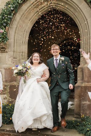 Happy couple exit church under shower of confetti while sharing joyful expressions.