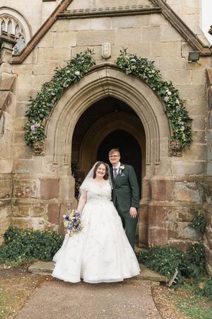 A couple stands in a Gothic stone church doorway decorated with a floral arch on their wedding day.