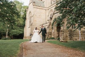 Newlyweds walking hand in hand along a path beside a historic stone church.