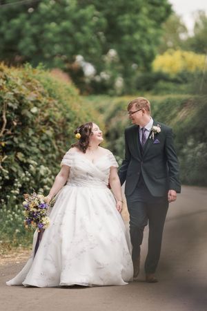 Newlyweds walk together on garden path surrounded by lush summer flowers.