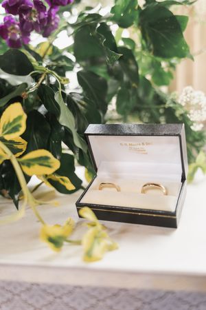 Two gold wedding rings displayed in an elegant black box with purple flowers in the background.