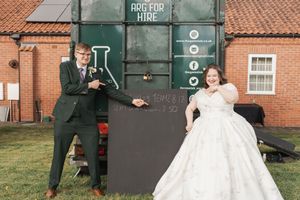 Wedding couple pose playfully in front of rustic brick building with vintage sign.