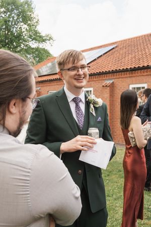 Wedding guest in dark green suit holding a drink and engaged in conversation during an outdoor reception.