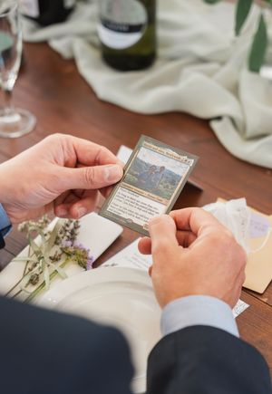 Hands holding a Magic: The Gathering card at an elegant table setting with dried flowers.