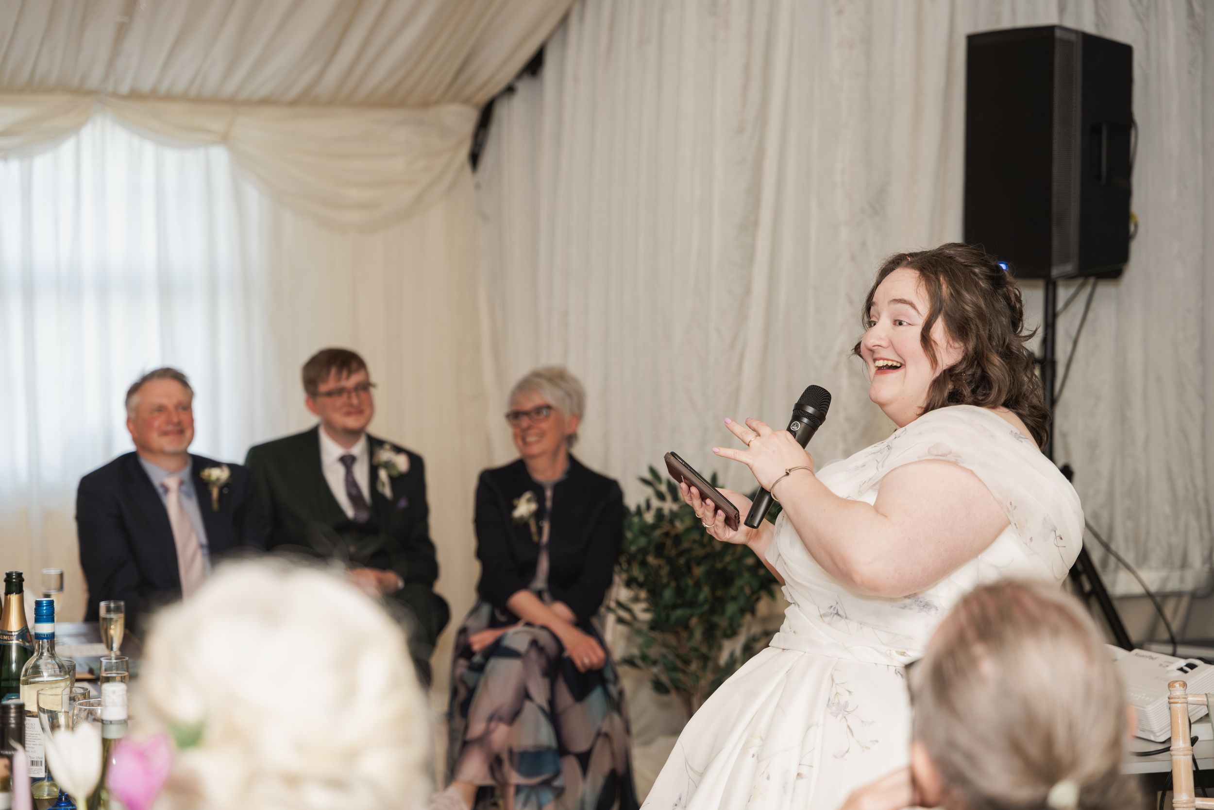 Person in white wedding dress giving a speech at a reception with guests seated at decorated tables.
