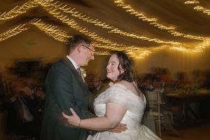 A couple dances together at a wedding reception under warm string lights and a tented ceiling.