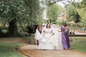 Bridesmaids in purple dresses assist the bride walking along a garden pathway.