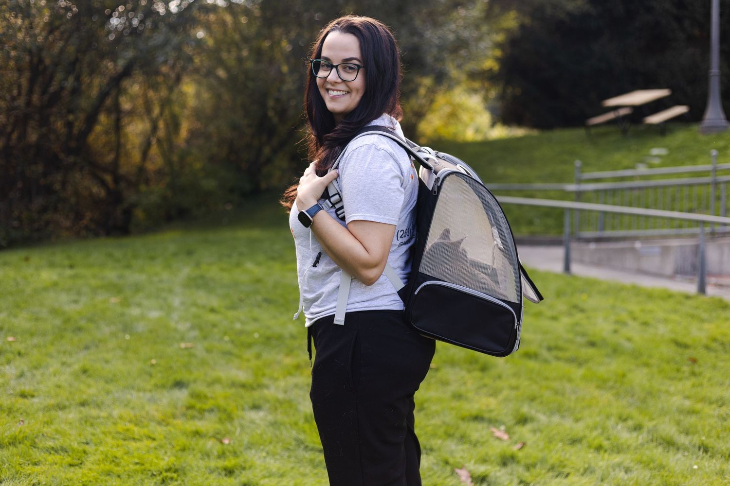 Student wearing glasses stands on grass with a backpack, smiling while holding backpack straps in an outdoor setting.