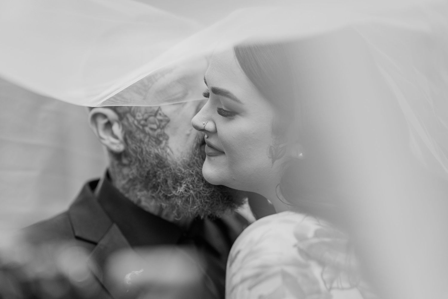 A tender black and white moment under a veil as a bearded groom and beautiful bride share an intimate embrace.