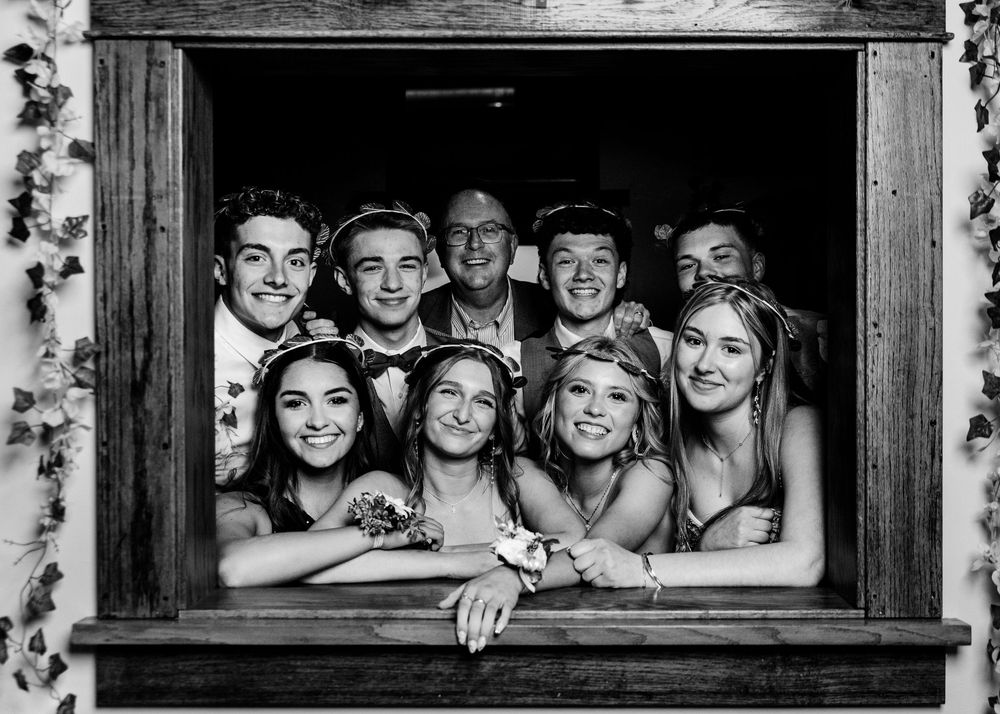A black and white photograph showing a group of people happily smiling while looking through a rustic wooden window frame.